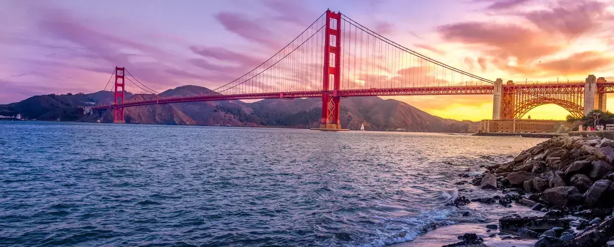 The 金门大桥 at sunset with a multicolored sky and the San Francisco Bay in the foreground.