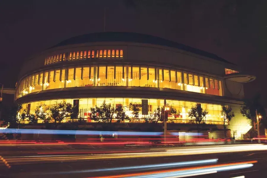 Exterior de la Sala Sinfónica Louise M. Davies por la noche con rayos de luz provenientes de vehículos que pasan.